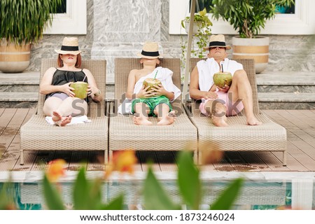 Father, mother and son covering faces with hats when relaxing on chaise-lounges with coconut cocktails in hands