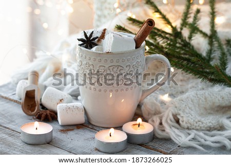 Winter hot drink. Cozy home composition with white mug with chocolate, marshmallow, cinnamon. Knitted mittens, christmas lights, wooden background. Festive holiday atmosphere, family spirit. Close up
