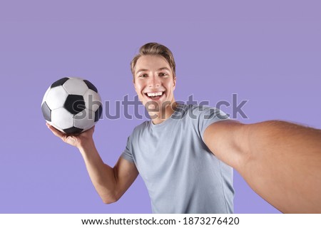 Smiling young man with soccer ball taking selfie on his mobile phone over violet studio background. Positive football fan taking photo of himself before championship, making good memories