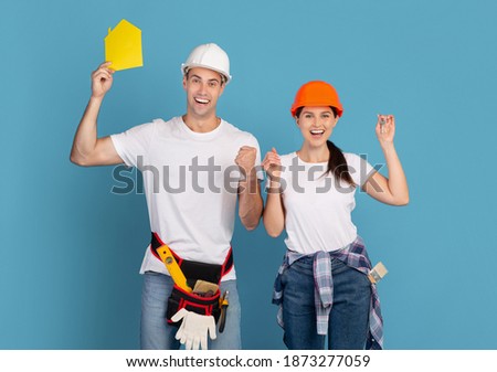 Joyful Millennial Couple In Hard Hats Holding Yellow Paper House Figure And Keys, Celebrating Ending Renovation, Raising Hands In Excitement, Standing Over Blue Studio Background, Copy Space