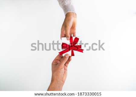 Beautiful holiday or Christmas background image of the hands of a Caucasian man and mixed race woman exchanging a small red ribbon wrapped white gift box on a white backdrop and copy space.