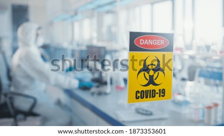 Danger Covid-19 Sign on the Glass Door of Virology and Medical Research Laboratory: Team of Scientists Wearing Masks and Coveralls Work on Covid-19 Cure, Vaccine, Developing Antibiotics, Drugs