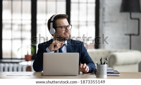 Dreamy businessman wearing headphones using laptop, sitting at desk in office, looking to aside, smiling employee executive dreaming, pondering future, listening to music after work done Royalty-Free Stock Photo #1873355149