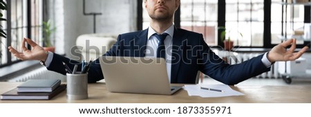 Horizontal banner calm businessman wearing suit meditating, practicing yoga in modern office, breathing deep, enjoying break, employee sitting at work desk with laptop, stress relief concept Royalty-Free Stock Photo #1873355971
