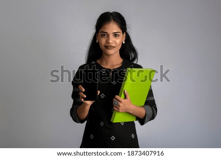 Pretty young girl holding book and using mobile on grey background