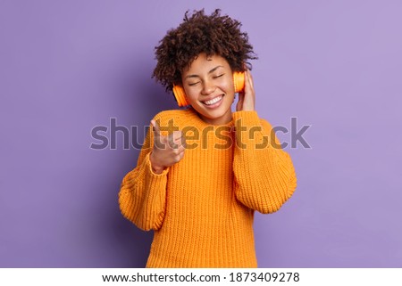 Optimistic curly haired woman makes excellent sign shows thumb up smiles pleasantly closes eyes rates song which she heard in stereo headphones wears orange jumper isolated over purple wall.