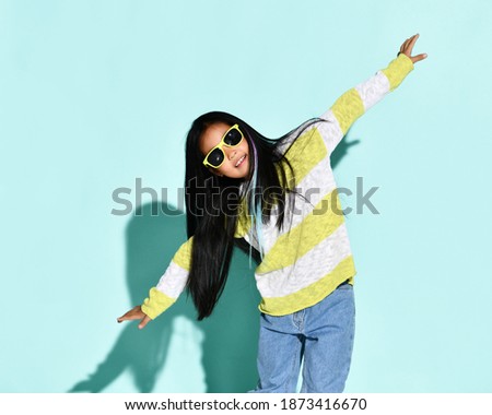 Portrait of stylish bright Korean girl posing over blue background with outstretched hands. A child in a bright sweater, jeans, sneakers and sunglasses. Kids fashion and style concept.