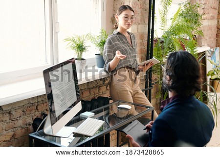 Young female mobile application developer with tablet having discussion with her male colleague drawing sketch in front of computer screen
