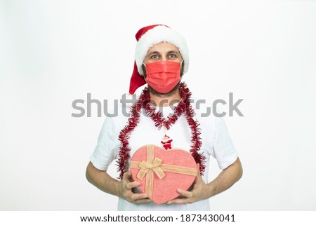 Man wearing a Santa hat. He has a red covid mask on his face. She is holding a heart-shaped gift box. isolated background.