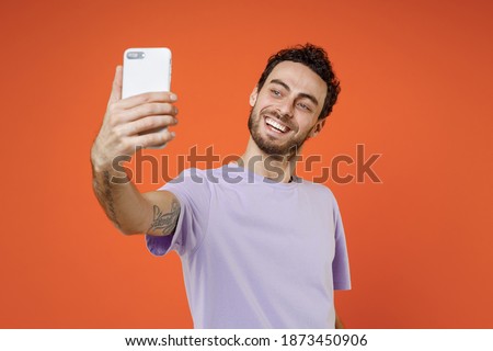 Cheerful smiling young bearded man 20s wearing casual basic violet t-shirt standing doing selfie shot on mobile phone isolated on bright orange color background studio portrait. Tattoo translate fun
