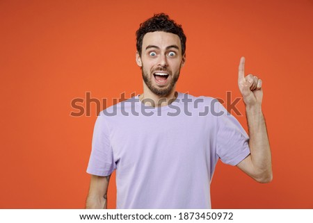 Excited amazed young bearded man 20s wearing basic casual violet t-shirt standing holding index finger up with great new idea looking camera isolated on bright orange color background studio portrait