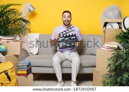 Cheerful owner man hold classic black film making clapperboard sits in living room on sofa at home household unpacking stuff rents flat isolated on yellow wall. Relocation moving in apartment concept Royalty-Free Stock Photo #1873450987