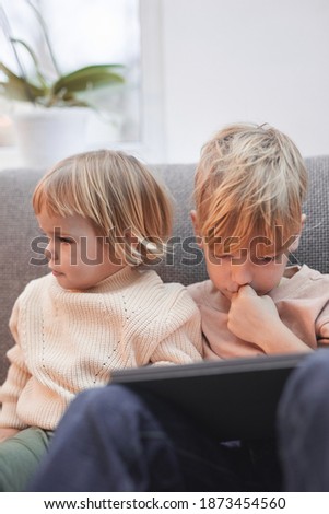 Vertical portrait of little boy and girl using digital tablet together while sitting on sofa, siblings and family concept