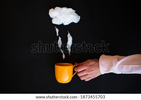 Hand holding Cup of coffee with steam and cloud made of cotton wool on black background with copy space. Flat lay. Weather concept