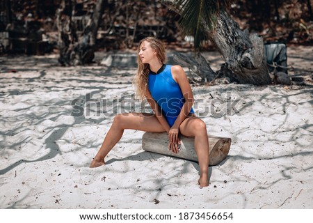 Young pretty blonde woman in a blue swimming suit sitting on the log of a palm tree. Summer vacation concept