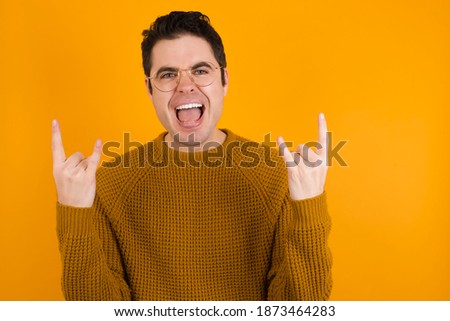 Born to rock this world. Joyful Young handsome Caucasian man wearing yellow sweater against orange wall screaming out loud and showing with raised arms horns or rock gesture.
