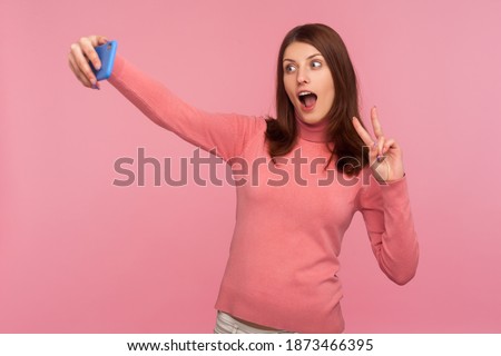 Funny female blogger with brown hair showing peace gesture and fooling face recording video or making photo on smartphone. Indoor studio shot isolated on pink background