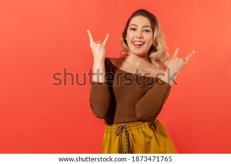 Yeah, that's awesome! Portrait of joyful woman in brown blouse, feeling crazy, showing devil horns, rock and roll hand gesture, delighted of success. Indoor studio shot isolated on red background