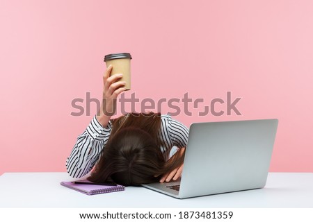 Sleepy bored woman office worker lying on table with laptop holding and showing paper coffee cup, feeling lack of energy sitting at workplace. Indoor studio shot isolated on pink background