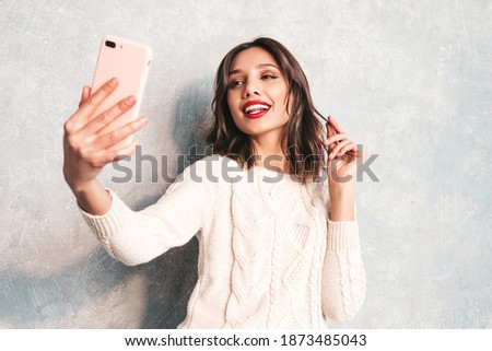 Young beautiful smiling woman looking at camera. Trendy female taking selfie photos at smartphone. Positive female with red lips taking selfie. Cheerful model posing near gray wall in studio 