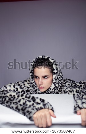 a woman in a leopard print hoodie lay down on a table with a white tablecloth, gray background, art portrait