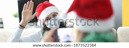 Friend take pictures of woman on phone in santa claus hat and tinsel. Sister pose and raise her hand up.
