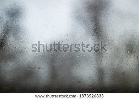 Selective focus and macro view of droplet of water, dew or stream on glass of window, and background of dim, foggy, hazy environment with blurry outer tree. 