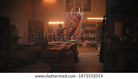 Back view of male artist painting portrait of black woman on easel against lamp during work in spacious workshop Royalty-Free Stock Photo #1873532014