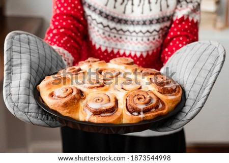 Woman in red holiday sweater holding glazed baked cinnabon buns with cinnamon on home backdrop. Christmas homemade bakery. Close up. 