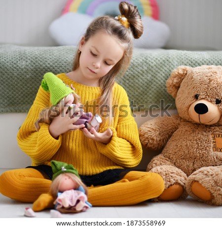 Pretty blonde small girl child in warm yellow knitted comfortable home sweater and tights sitting on floor and playing with doll and teddy bear at home. Happy childhood, games, leisure, hobby