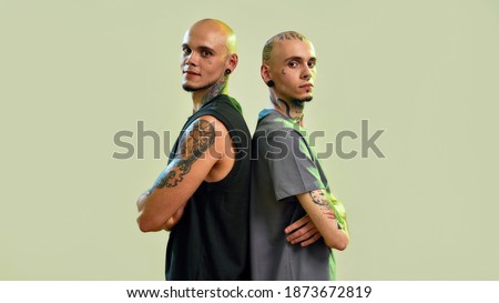 Portrait of two young men, twin brothers with tattoos and piercings looking at camera, posing together, standing back to back isolated over light green background. Web Banner