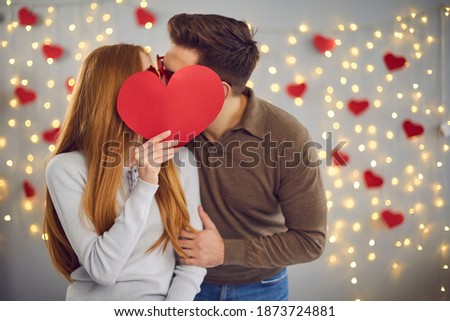 Love in the air. Unknown young man and woman kissing hiding behind a big red paper heart they are holding in hand. Valentine's day, kissing, love and youth concept. Banner. Place for text. Ad layout.