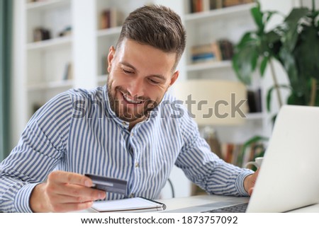 Smiling man sitting in office and pays by credit card with his laptop.