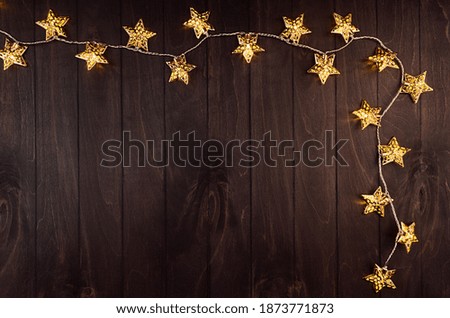 Bright festive background with golden stars glowing of electric lights on elegant dark brown board, frame, top view.