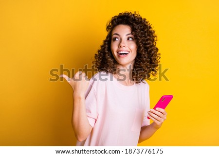 Photo portrait of curly dreamy girl holding mobile phone showing at copyspace smiling isolated on bright yellow color background
