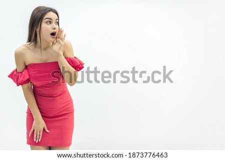 Copyspace photo of beautiful young woman in red dress, wispering some secret to the camera, keeping hand near mouth over white background.