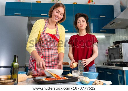 Young Caucasian women prepare food together in the kitchen. They spread the sauce on the pizza and grate the cheese. The concept of home-cooked food and LGBT relationships