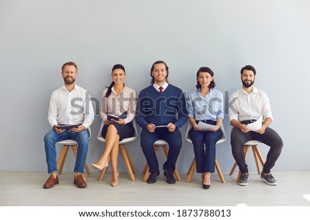 Group of smiling candidates job seekers sitting on chairs in row with resumes in hand and waiting in line for interview invitation turn in office of modern company. Office career seekers concept Royalty-Free Stock Photo #1873788013