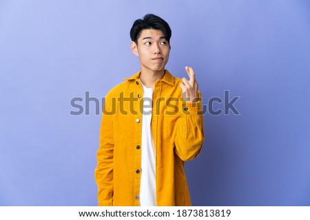 Young Chinese man isolated on purple background with fingers crossing and wishing the best