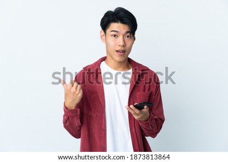 Young Chinese man isolated on white background using mobile phone and pointing to the lateral