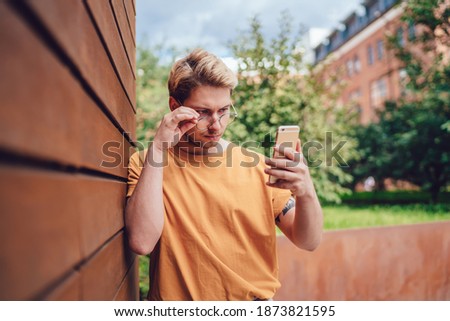 Serious adult freelancer in glasses and casual orange shirt focusing on screen and trying to solve problem while standing leaning on brown striped wall and using smartphone against green trees in city