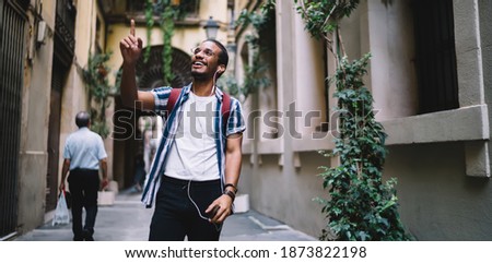 Glad young ethnic male traveler having sightseeing with audioguide on smartphone with earphones and pointing up by finger on narrow street with plants in old town Royalty-Free Stock Photo #1873822198