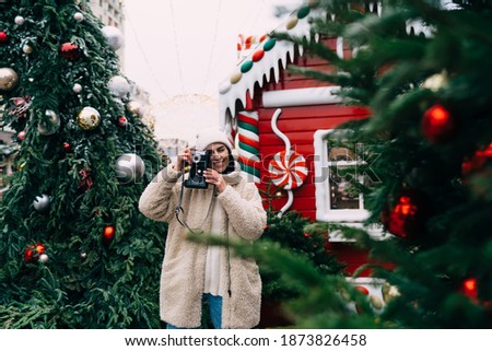 Joyful female photographer in warm winter clothes shooting pictures with photo camera while spending winter holidays at wonderful Christmas market