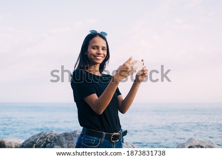 Latin blogger smiling at camera while using smartphone application for clicking photo images at coastline, happy female millennial holding cellular gadget and posing during summer vacations