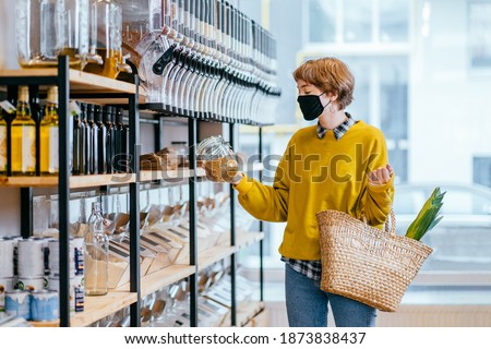 Shopping, food, sale, consumerism concept. Blond short haired woman in face protective mask holds glass jar with soya, buying at grocery store Reusable wicker basket for shopping. Zero waste concept. Royalty-Free Stock Photo #1873838437