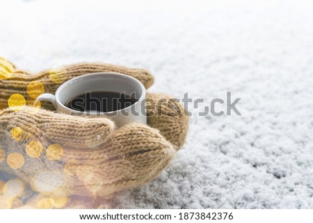 Hands in woolen knitted gloves holding cup of warm black coffee outdoors on snowy background. Top view of hands and coffee mug. Happy, frosty winter morning and hygge concept