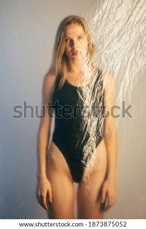 Beauty care. Defocused female body. Anti cellulite treatment. Weight loss. Slim woman in black bodysuit standing behind wrinkled texture polyethylene film isolated on gray background out of focus.