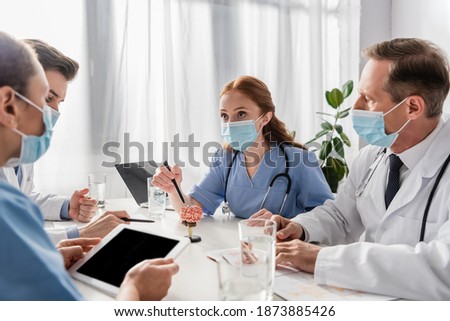 Multicultural nurses and doctors working while sitting at workplace with devices, papers and glasses of water on blurred foreground