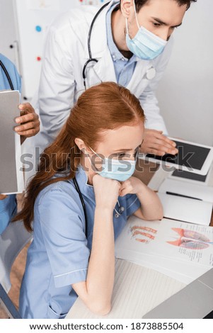 Nurse in medical mask sitting at workplace near multicultural colleagues on blurred background