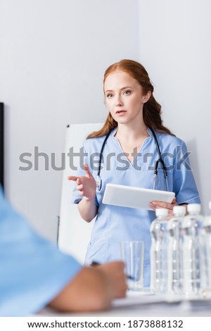Nurse pointing with finger while holding digital tablet near colleagues and bottles of water on blurred foreground